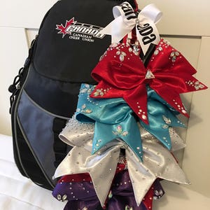 May include: A black backpack with the logo "Canada Canadian Cheer Union" and a white ribbon with the text "Canada" attached to it. The backpack is decorated with five large, sparkly cheer bows in red, teal, silver, and purple.