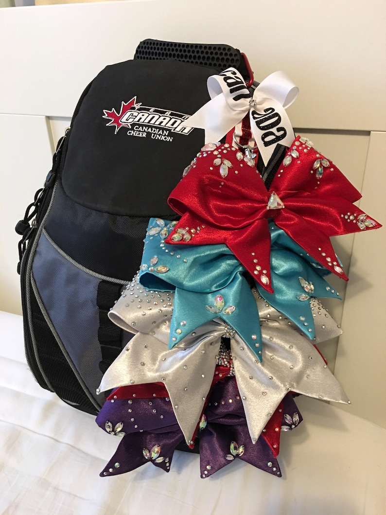 May include: A black backpack with the logo "Canada Canadian Cheer Union" and a white ribbon with the text "Canada" attached to it. The backpack is decorated with five large, sparkly cheer bows in red, teal, silver, and purple.