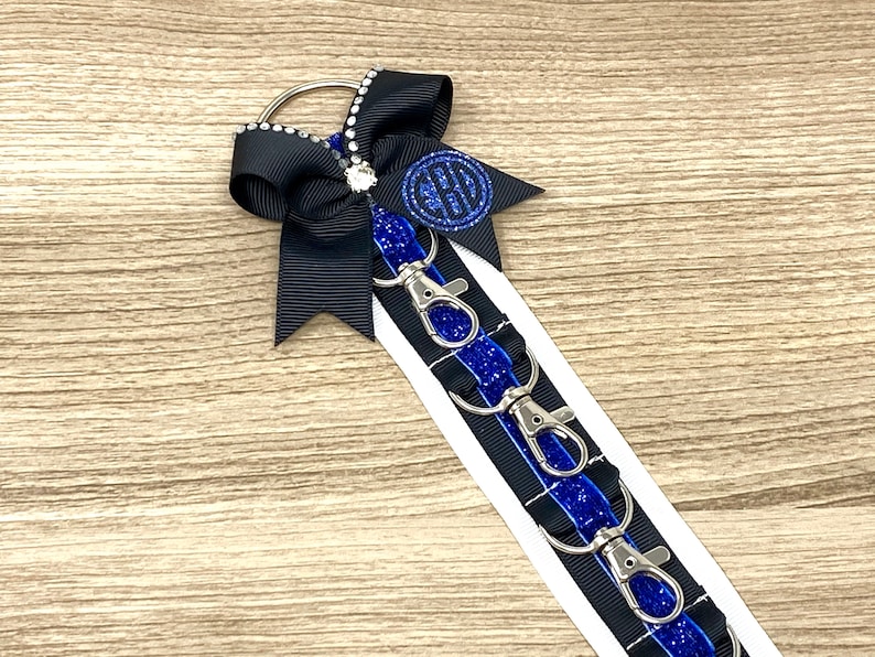 May include: A black and blue key chain with a black bow and a silver charm with a monogram. The key chain has five silver key rings.