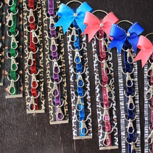 May include: A set of eight keychains with different coloured ribbons and animal print fabric. Each keychain has multiple silver hooks. The ribbons are gold, green, red, purple, blue, pink, black and white.