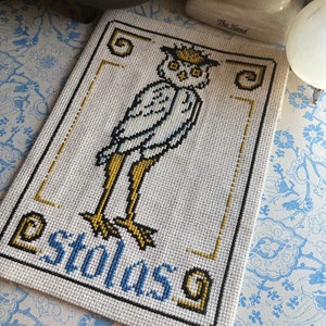 May include: A white owl with yellow feet and a crown on its head is embroidered on a white canvas. The owl is surrounded by a gold frame and the word "Stolas" is written in blue and black letters below the owl.