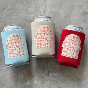 May include: Three beverage can coolers in blue, beige, and red. Each cooler has a white graphic with a cowboy hat, hot dog, baby bottle, and burger, with the text "Baby-Q" in a script font.