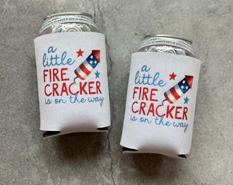 Puede incluir: Dos enfriadores de latas blancos con el texto "a little FIRE CRACKER is on the way" en azul y rojo. Cada enfriador tiene un gráfico de un petardo con estrellas y rayas rojas, blancas y azules.