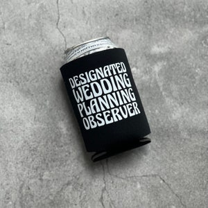 May include: Black neoprene can cooler with white text that reads "Designated Wedding Planning Observer".