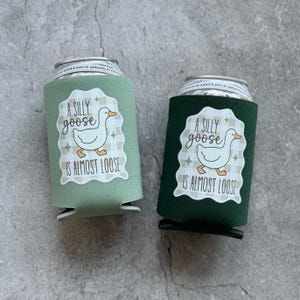 Op de afbeelding: Twee drankblikjeskoelers, &eacute;&eacute;n saliegroen en &eacute;&eacute;n donkergroen. Elk heeft een wit label met een cartoon gans en de tekst "A SILLY goose IS ALMOST LOOSE". De achtergrond is een grijs, getextureerd oppervlak.