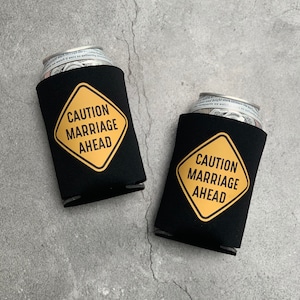 May include: Two black foam beverage insulators with a yellow caution sign that reads "Caution Marriage Ahead".