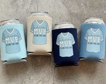 May include: Four beverage can coolers in shades of blue, tan, and navy. Each cooler features a light blue t-shirt design with the text "a little MVP IS ON THE WAY". The coolers are designed to keep beverages cold.