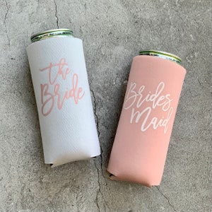 May include: Two white and pink can coolers with the text "the bride" and "bridesmaid" in pink lettering.