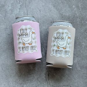 Op de afbeelding: Twee drankblikjeskoelers, &eacute;&eacute;n roze en &eacute;&eacute;n beige, elk met een witte sticker met een cartoon gans en de tekst "A Silly Goose Is Almost Loose". De koelers zijn ontworpen om drankjes koud te houden.
