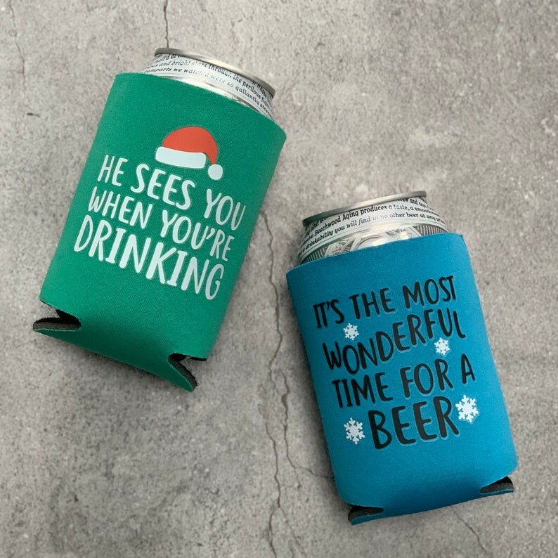 May include: Two green and blue insulated beverage holders with Christmas-themed sayings. The green holder says "He sees you when you're drinking" with a red Santa hat graphic. The blue holder says "It's the most wonderful time for a *beer*" with snowflake graphics.