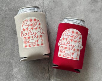Puede incluir: Dos fundas para latas, una beige y otra roja, cada una con un diseño "Baby-Q". El diseño incluye un sombrero de vaquero, un perrito caliente, una hamburguesa, un biberón y un chupete, todo sobre un fondo a cuadros rojos y blancos.