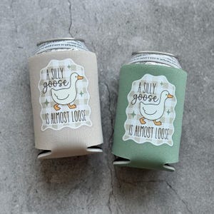 Op de afbeelding: Twee drankblikjeskoelers, &eacute;&eacute;n beige en &eacute;&eacute;n groen, elk met een wit etiket met een ganzenillustratie en de tekst "A SILLY goose IS ALMOST LOOSE". De koelers zijn ontworpen om drankjes koud te houden.