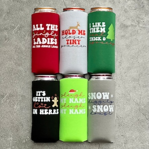 May include: A collection of six beverage can coolers in various colors, each with a different holiday-themed phrase. The coolers are red, white, green, black, lime green, and gray. Phrases include "All the Jingle Ladies" and "Hold me closer tiny prancer."