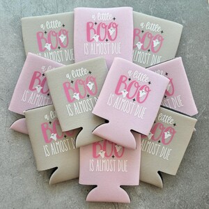 May include: A collection of pink and tan can coolers with the phrase "a little BOO is ALMOST DUE" printed in pink and white. Each cooler features a ghost graphic, perfect for Halloween or baby showers.