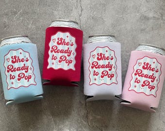 Puede incluir: Cuatro enfriadores de latas de espuma en azul claro, rojo, lavanda y rosa claro. Cada enfriador presenta una etiqueta festoneada blanca con rayas azul claro y el texto rojo "She's Ready to Pop" con corazones y estrellas.