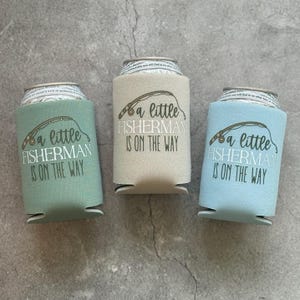 May include: Three different coloured can coolers with the text "a little fisherman is on the way" printed on them. The can coolers are green, beige, and blue.