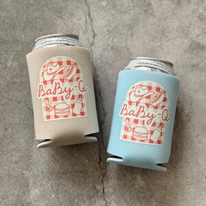 May include: Two beverage can coolers, one tan and one light blue, each featuring a red and white checkered design with the text "Baby-Q" and illustrations of a cowboy hat, hot dog, baby bottle, and burger.