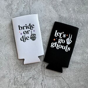 May include: Two white and black beverage coolers with Halloween-themed text. The white cooler says "bride or die" with a skeleton hand giving the middle finger. The black cooler says "let's go ghouls" with a skeleton hand giving the peace sign.
