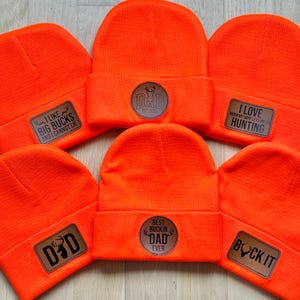 May include: Six orange knit beanies with different brown leather patches. The patches have various hunting-themed sayings and images, including "I like big bucks and I cannot lie", "Best Buckin' Dad Ever", "I love when my wife lets me go hunting", and "Dad est. 2025".