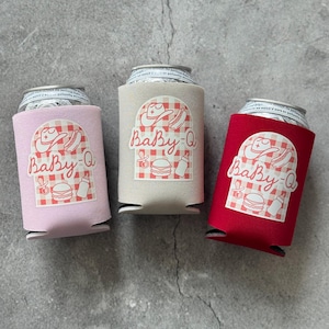 May include: Three can coolers in pink, tan, and red. Each cooler has a white graphic with a cowboy hat, hot dog, baby bottle, and burger, with the text "Baby-Q" in a script font. The coolers are on a gray surface.
