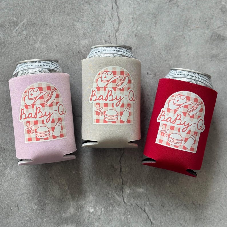May include: Three can coolers in pink, tan, and red. Each cooler has a white graphic with a cowboy hat, hot dog, baby bottle, and burger, with the text "Baby-Q" in a script font. The coolers are on a gray surface.