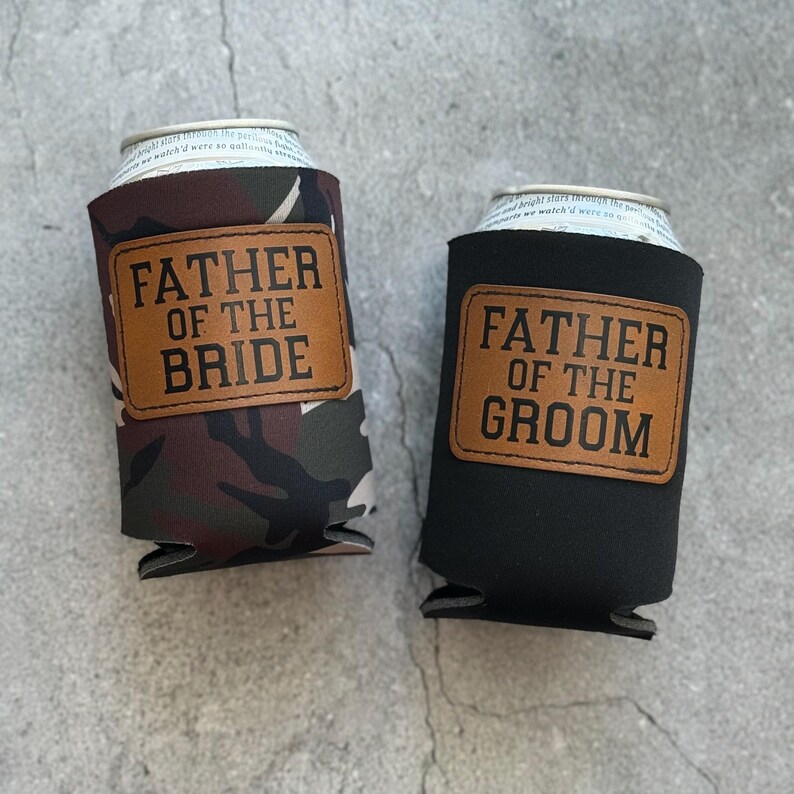 May include: Two can coolers, one with a camouflage pattern and the other black. Both have a brown leather patch with the words "FATHER OF THE BRIDE" and "FATHER OF THE GROOM" in capital letters.