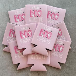 May include: A collection of pink can coolers with the phrase "a little BOO is almost due" printed in white and pink. Each cooler features a ghost graphic. The coolers are designed for a Halloween or baby shower theme.