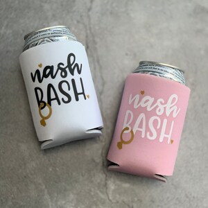 May include: Two white foam can coolers with the text "nash bash" in black lettering and a gold ring symbol. One cooler is white and the other is pink.