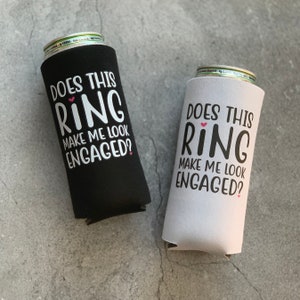 May include: Two black and white can coolers with the text "Does this ring make me look engaged?" in white with a pink heart above the word "ring".