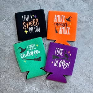 May include: Four colorful Halloween-themed beverage insulators with different sayings. The sayings are "I put a spell on you", "I smell children", "Amuck amuck amuck", and "Come, we fly!"