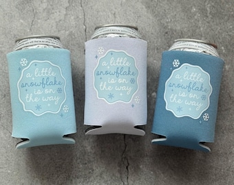 May include: Three beverage can coolers in shades of blue and gray. Each cooler has a white label with the text "a little snowflake is on the way" and snowflake graphics. The coolers are designed to keep drinks cold.