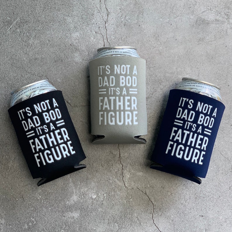May include: Three different coloured can coolers with the text "It's not a dad bod = It's a father figure" printed on them. The can coolers are black, grey, and navy blue.
