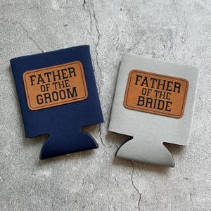May include: Two beverage insulators, one navy blue and one gray. Each has a brown leatherette patch with the words "FATHER OF THE GROOM" and "FATHER OF THE BRIDE" in bold, black letters. The insulators are designed to hold a standard can or bottle.