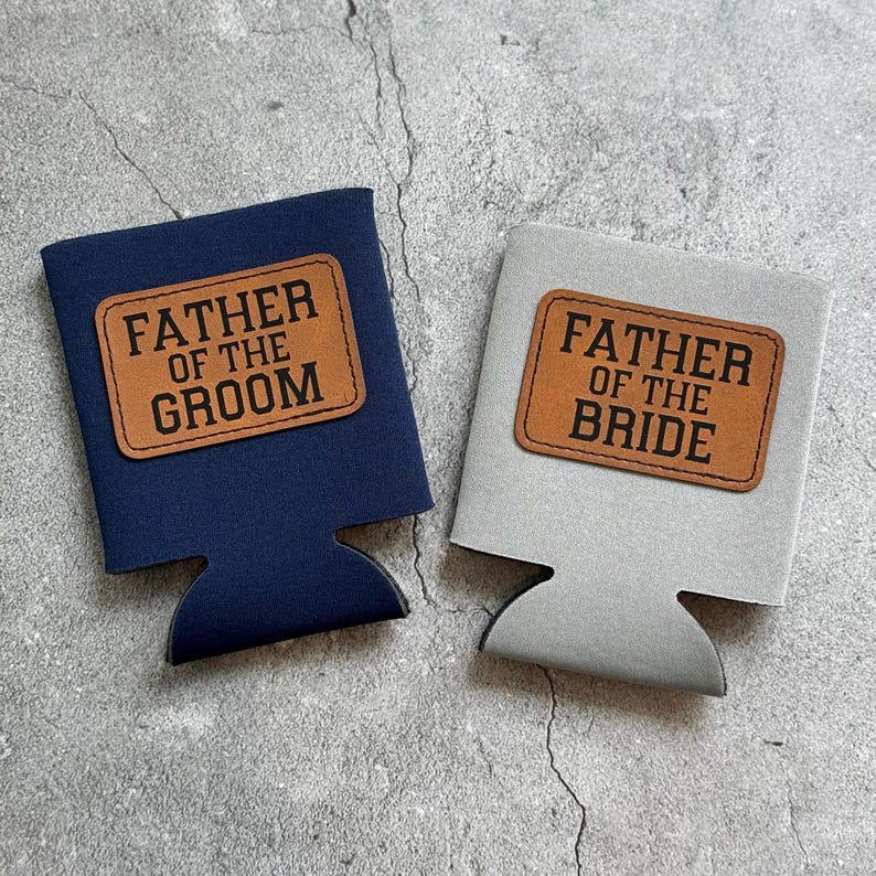 May include: Two beverage insulators, one navy blue and one gray. Each has a brown leatherette patch with the words "FATHER OF THE GROOM" and "FATHER OF THE BRIDE" in bold, black letters. The insulators are designed to hold a standard can or bottle.