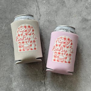 May include: Two can coolers, one tan and one pink, each holding a can. The coolers have a "Baby-Q" design with a cowboy hat, hot dog, baby bottle, and burger on a red and white checkered background.