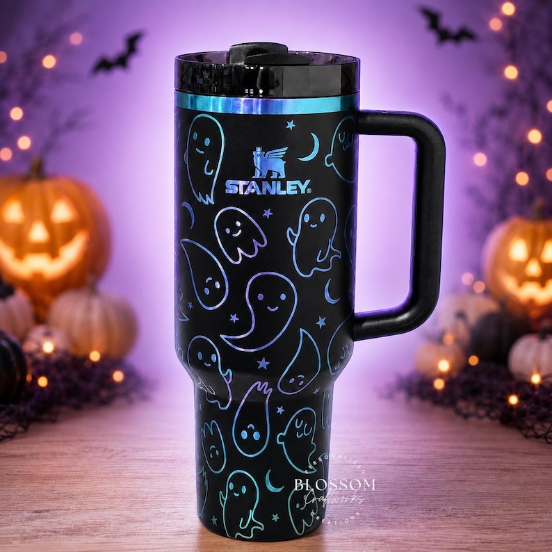 May include: A black Stanley tumbler with a handle and a lid, featuring a repeating pattern of iridescent blue and purple ghosts, stars, and crescent moons. The Stanley logo is printed in the center in a matching iridescent blue and purple finish.