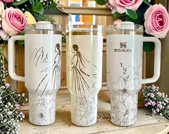 May include: Three white Stanley tumblers with floral and bride-themed designs. One tumbler has the word "Mrs" in script, another features a bride illustration, and the third has the Stanley logo. Each tumbler has a handle and a clear lid.