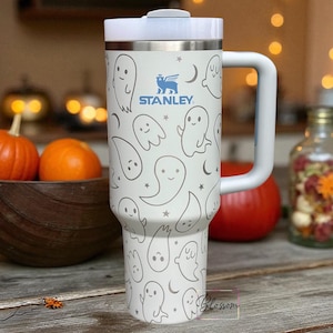 May include: A white Stanley tumbler featuring a repeating pattern of grey ghosts, stars, and crescent moons. The insulated cup has a large side handle, a stainless steel rim, and a white lid. The Stanley logo is printed in blue on the front.