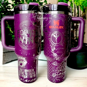 May include: Purple stainless steel travel mug with a black lid and handle. The mug has a silver design with a dragon and the text "Our Wing" and "I am the storm".