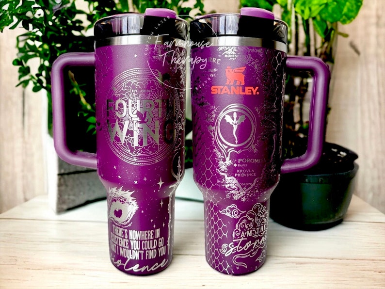 May include: Purple stainless steel travel mug with a black lid and handle. The mug has a silver design with a dragon and the text "Our Wing" and "I am the storm".