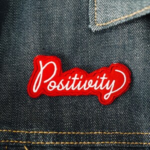 Könnte beinhalten: Roter Stoff-Patch mit weißer Aufschrift "Positively" auf Jeans-Hintergrund.