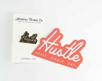 May include: A gold enamel pin with black lettering that reads "Hustle Every Single Day." The pin is on a white card with the text "Mindful Things Co. Gear for the Inspired Journey." There is also a red sticker with white lettering that reads "Hustle Every Single Day."