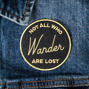 May include: A black and gold embroidered patch with the text "Not all who wander are lost" on a denim background.