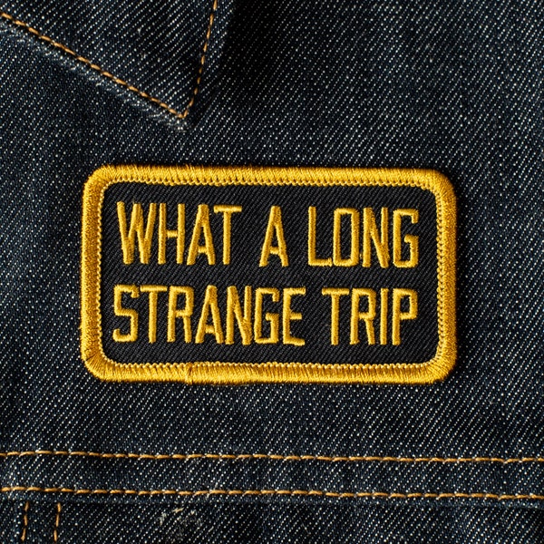 May include: A black rectangular patch with gold embroidered text that reads "WHAT A LONG STRANGE TRIP." The patch has a gold border and is sewn onto a dark blue denim fabric. The denim has visible stitching.