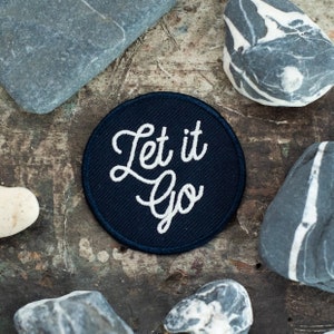 Puede incluir: Un parche redondo de color azul oscuro con bordado blanco que dice "Let it go". El parche está rodeado de piedras grises.