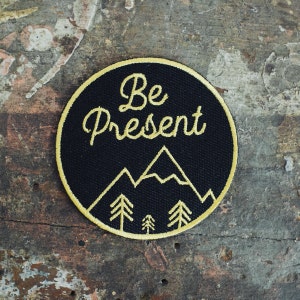 May include: Black embroidered patch with yellow stitching. The patch features a mountain range and the text "Be Present".