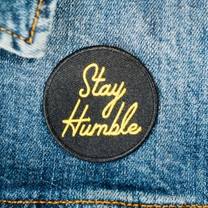 Peut inclure: Un écusson rond noir avec l'inscription "Stay Humble" brodée en jaune. L'écusson est fixé sur une veste en jean bleue, avec la texture du tissu et les coutures visibles.