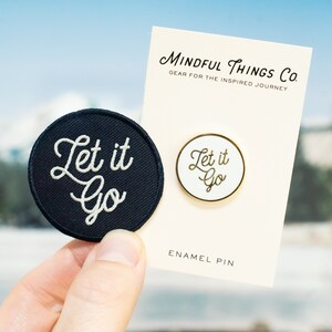 May include: A black and white embroidered patch and a gold enamel pin, both with the text "Let it Go" in a cursive font. The patch and pin are on a white card with the text "Mindful Things Co. Gear for the Inspired Journey" and "Enamel Pin".