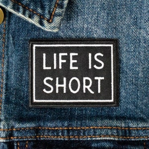 Pode incluir: Um remendo preto com costuras brancas sobre um fundo de denim azul. O remendo diz "Life is Short" em letras brancas.