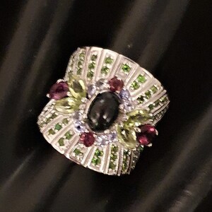May include: A silver ring featuring a black gemstone surrounded by green, purple, and white stones.